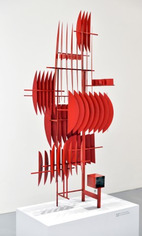 Abstrakcyjna rzeźba z pomalowanego na czerwono metalu. Pionowe i poziome pręty tworzą nieregularny przestrzenny stelaż. Na nim w różnych miejscach przyspawane cienkie blaszki w formie wycinków koła, ustawione blisko siebie i jakby zmultiplikowane.