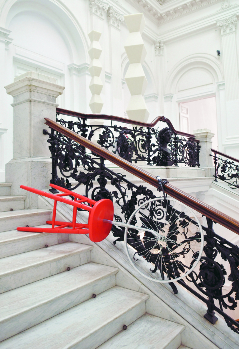 Na zdjęciu białe, dekoracyjne wnętrze klatki schodowej Zachęty; na schodach przewrócony czerwony stołek barowy z widelcem rowerowym wbitym w siedzisko; na widelcu koło przypięte blokadą do poręczy schodów.
