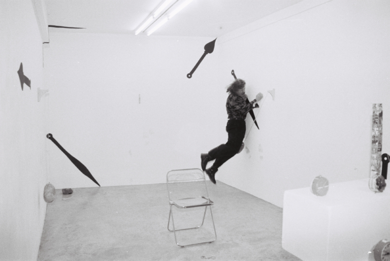 Czarno-białe zdjęcie: jasne pomieszczenie z krzesłem na środku oraz czarnymi symbolami na ścianach – strzałką i wskazówkami; w prawym rogu mężczyzna z długimi włosami, uchwycony w podskoku, jakby ciśnięty przez niewidoczna siłę o ścianę.