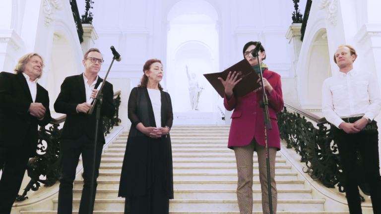 Zdjęcie: jasna, dekoracyjna klatka schodowa Zachęty. Na schodach, za dwoma mikrofonami stoi trzech mężczyzn i dwie kobiety. Jedna z kobiet trzyma przed sobą otwartą brązową okładkę z godłem – przemawia.