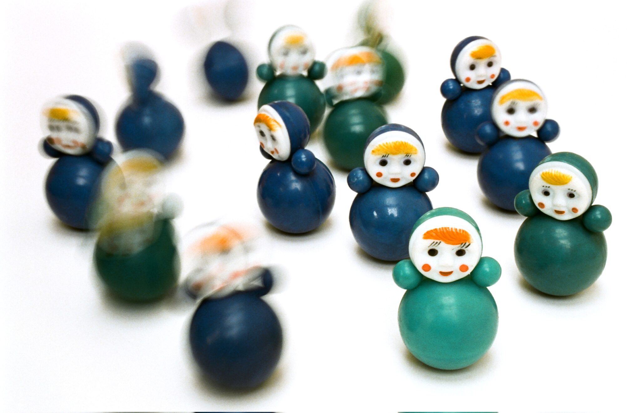 Na zdjęciu 11 niebieskich i zielonych zabawek typu wańka-wstańka, czyli kiwających się na boki; każda złożona z 4 kulek: korpusu, głowy przypominającej uśmiechnięty księżyc w pełni i dwóch rączek; dwie zabawki uchwycone w ruchu, rozmazane.