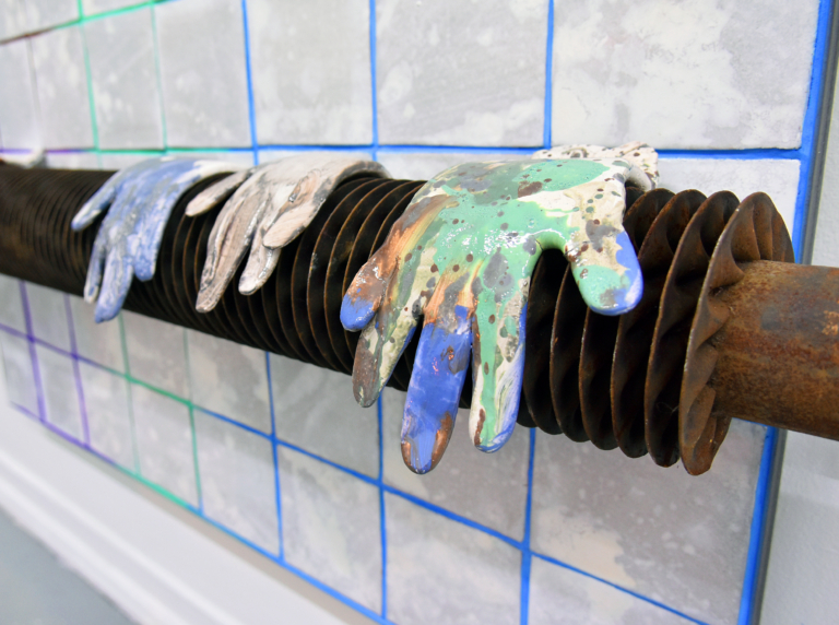 Zdjęcie: na tle ściany, wyłożonej jasnymi kafelkami, kaloryfer starego typu: w kształcie żebrowanej rury; na kaloryferze wiszą robocze rękawice ochlapane kolorową farbą; przeważa zielony i niebieski, który koresponduje z niebieską fugą.