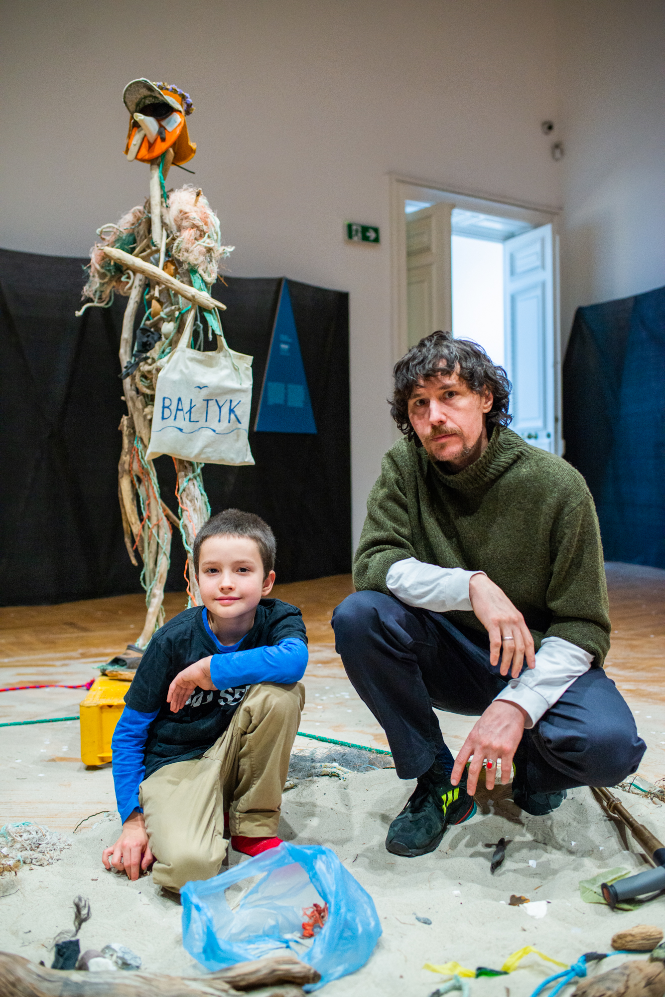We wnętrzu, na parkiecie, wyspa z piachu i odpadów; na niej przykucnęli: krótko ostrzyżony, uśmiechnięty chłopiec, wiek ok. 10 lat i starszy od niego mężczyzna z wąsami i brodą; w tle postać z konarów i śmieci, w klapkach, z torbą podpisaną: Bałtyk.