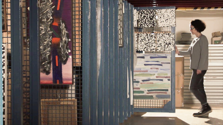 Zdjęcie wnętrza magazynu. Z lewej strony jedna za drugą wiszą niebieskie metalowe kraty o drobnych oczkach. Na pierwszej wisi obraz. W głębi kobieta w szarym swetrze wysuwa jedną z krat, na której wiszą abstrakcyjne obrazy.