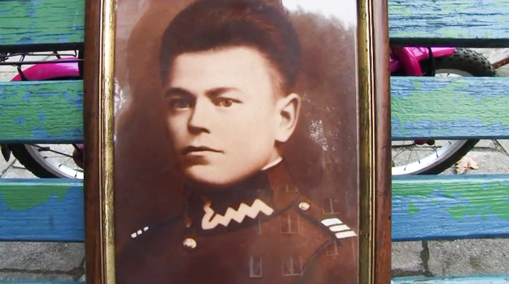 Kadr filmu: stare zdjęcie portretowe młodego mężczyzny w mundurze. Zdjęcie w odcieniach brązu. Wisi na zielononiebieskich deskach, spomiędzy których wyłania się różowy rower.