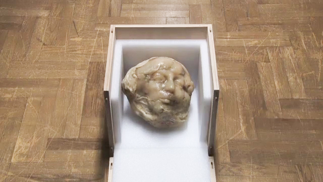 Kadr filmu: na podłodze z klepek stoi drewniana skrzynka bez przedniej ściany; wyłożona jest białą pianką. W środku leży rzeźba kobiecej głowy z tworzywa sztucznego w cielistym kolorze. Głowa jest zniekształcona.