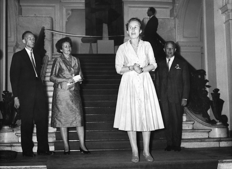 Czarno-białe zdjęcie: na wysokich schodach stoi szczupła kobieta w białej sukience. Trzyma się za dłonie. W tle, z prawej strony mężczyzna w garniturze.