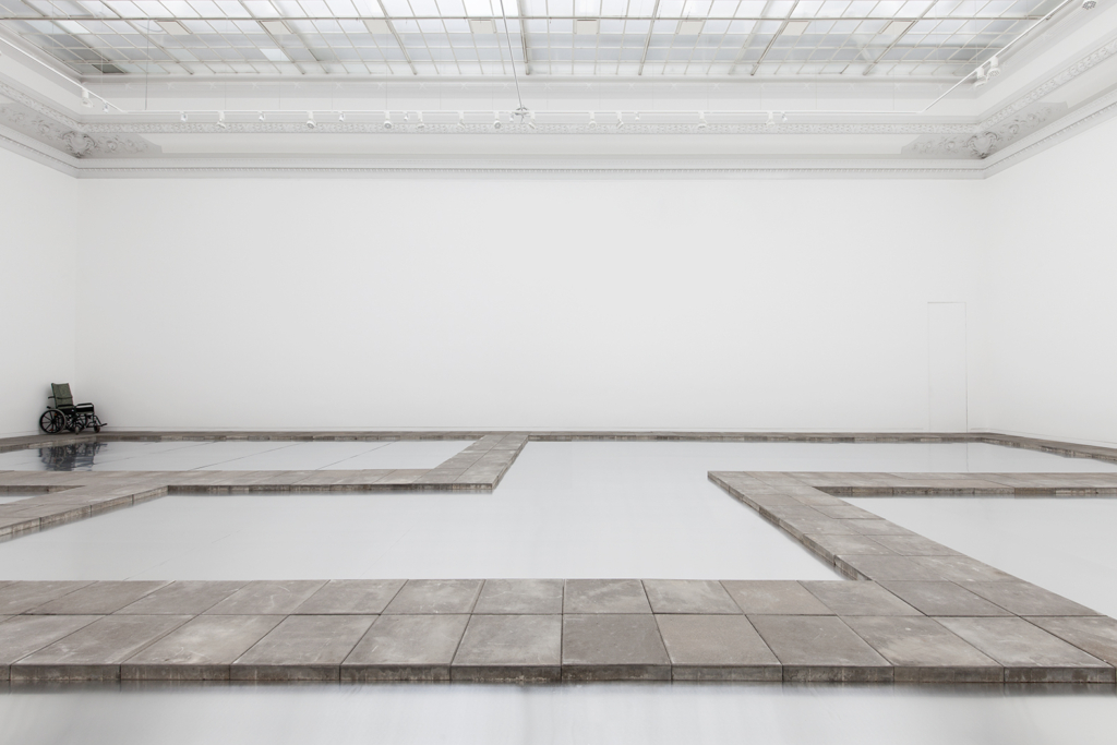 Zdjęcie: wnętrze sali o białych ścianach i lekko niebieskiej posadzce, na podłodze ułożone w nieskomplikowany wzór ścieżki z szarych, betonowych płyt chodnikowych; w rogu sali, na najdalszym planie fotografii, wózek ortopedyczny.