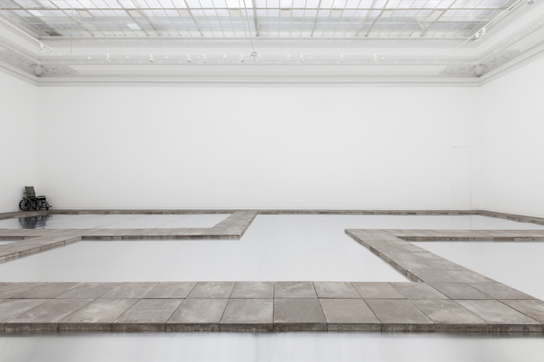 Zdjęcie: wnętrze sali o białych ścianach i lekko niebieskiej posadzce, na podłodze ułożone w nieskomplikowany wzór ścieżki z szarych, betonowych płyt chodnikowych; w rogu sali, na najdalszym planie fotografii, wózek ortopedyczny.