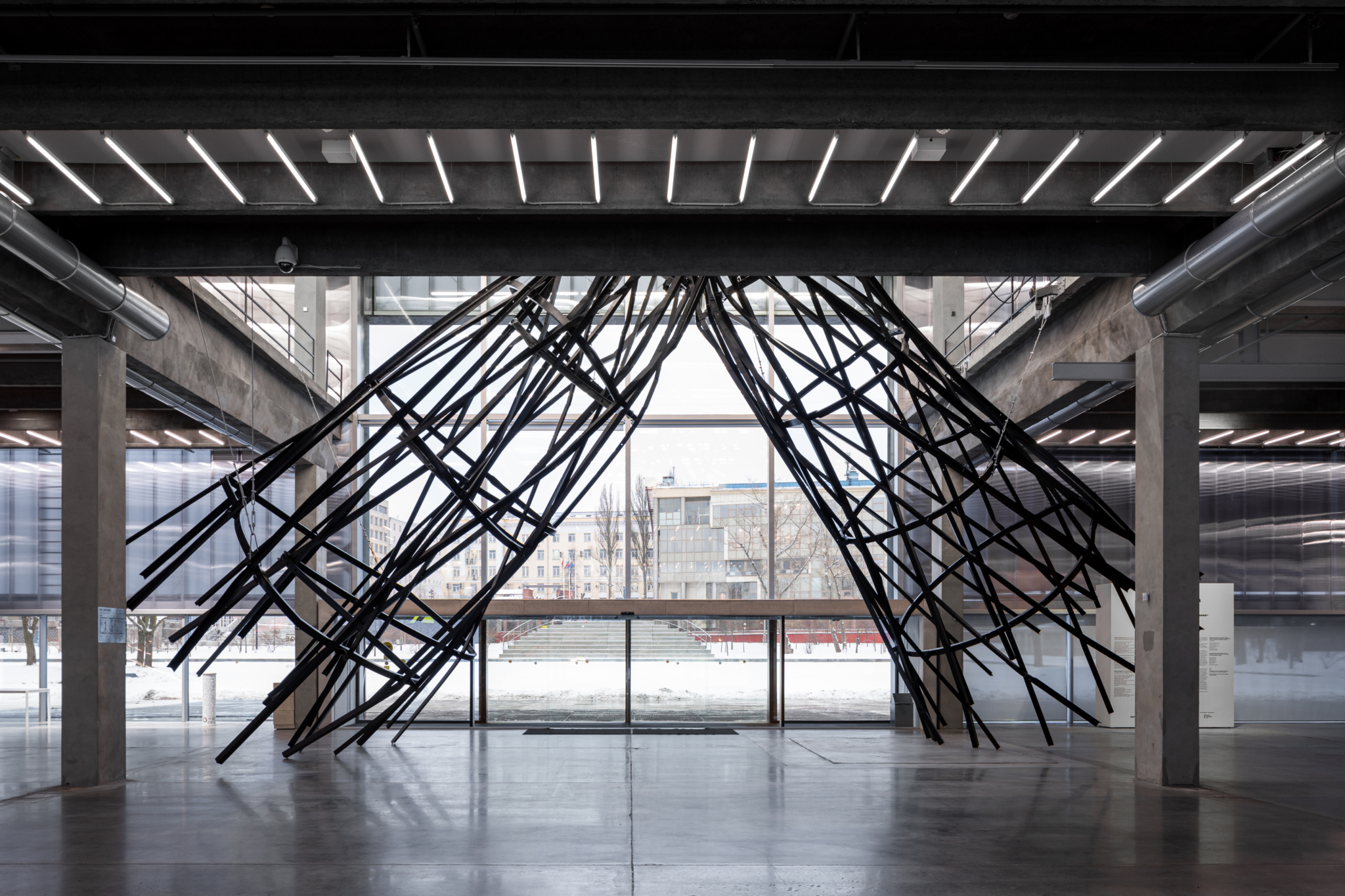 Przeszkola przestrzeń wystawiennicza. Na środku wielkoformatowa rzeźba ze stalowych prętów przypominająca wieżę nadawczą zgiętą w połowie