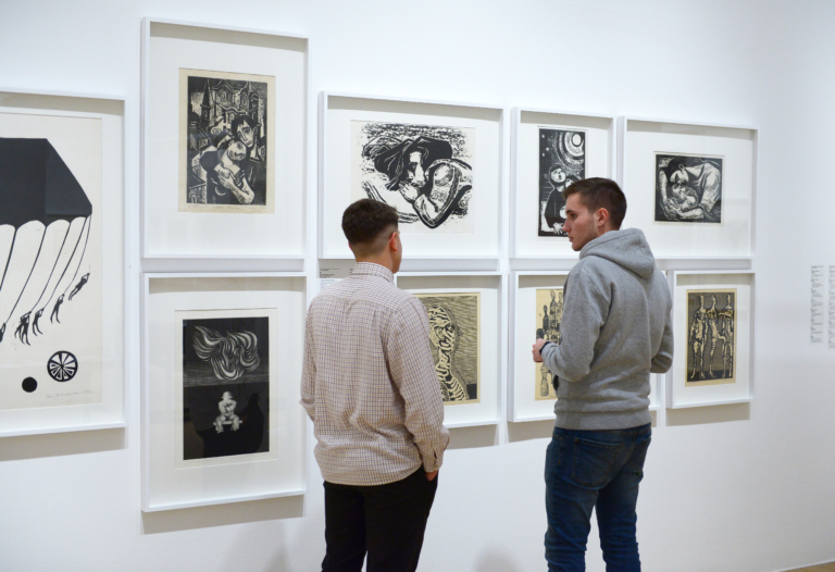 Dwóch, młodych mężczyzn stoi na tle ściany, na której wisi 8 grafik z kolekcji Zachęty