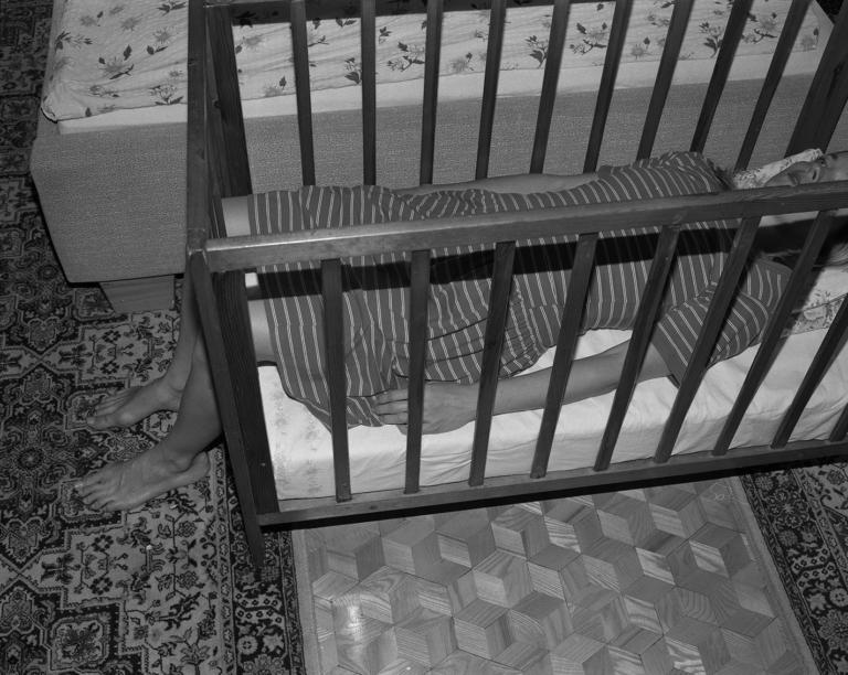 Czarno-białe zdjęcie. W łóżeczku dla dziecka leży dorosła kobieta, nogi wystają jej poza łóżko.