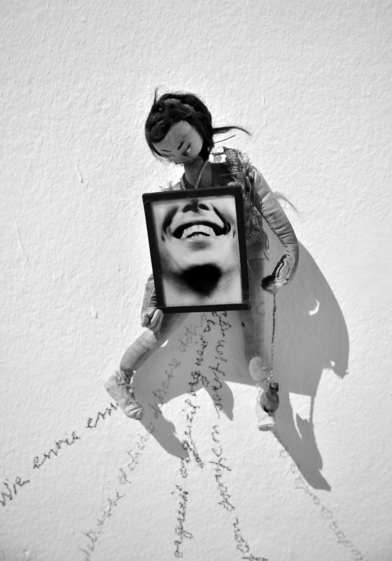 Czarno-białe zdjęcie. Na ścianie wisi lalka, która trzyma zdjęcie twarzy kobiety