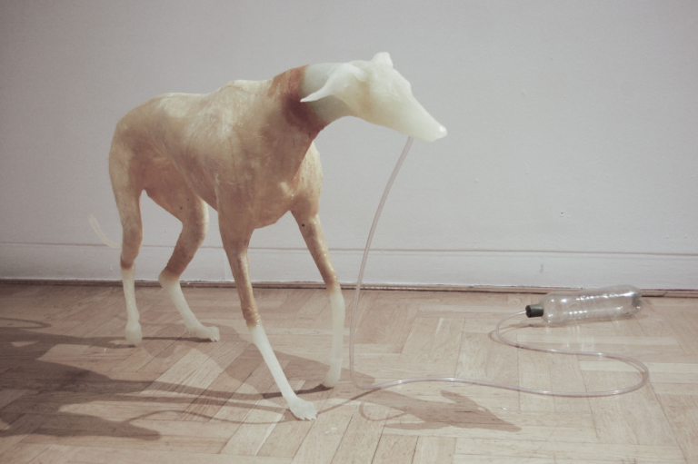 Figura psa z jasnego wosku, przypominająca charta. Pies trzyma w pysku plastikową rurkę połączoną z plastikową butelką