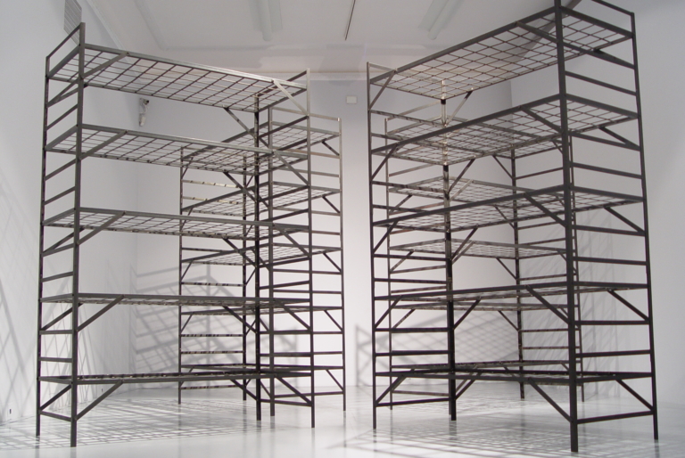 Przestrzeń ekspozycyjna w której stoją trzy metalowe, wielopiętrowe łóżka, przypominające więzienne lub obozowe prycze