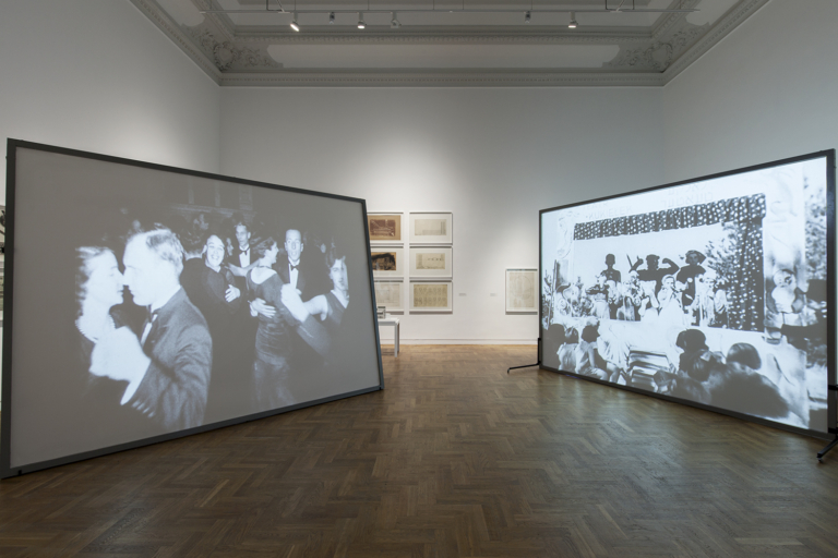 Zdjęcie: widok ekspozycji wystawy. Na środku sali stoją dwa, duże ekrany do projekcji filmowej. Na obu wyświetlane są czarno-białe filmy