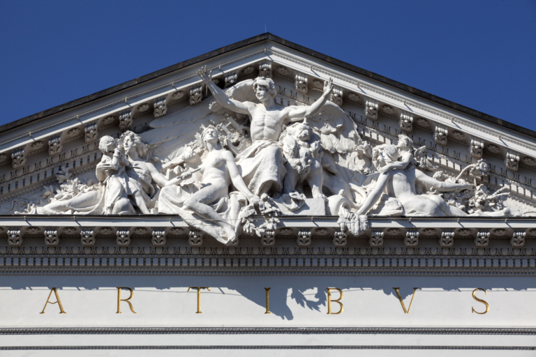 Opis zdjęcia: zdjęcie przedstawia zbliżenie na fasadę Zachęty – klasycyzujący budynek z 1900 roku, z tympanonem i napisem ARTIBUS (sztukom).