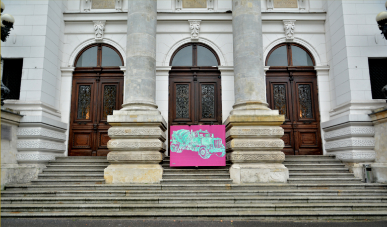 5fcf7ddd57e16 Zdjęcie: schody prowadzące do wejścia głównego Zachęty; trzy pary drzwi przedzielone dwoma pilastrami. Przed środkowymi drzwiami stoi kobieta trzymająca obraz – turkusową betoniarkę na ciemno różowym tle
