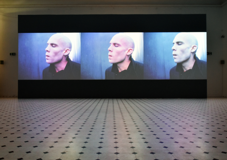 Zdjęcie: na ścianie sali wystawowej wyświetlana jest trój-kanałowa projekcja. Na ekranie jest zbliżenie na twarz młodego mężczyzny z wygoloną głową
