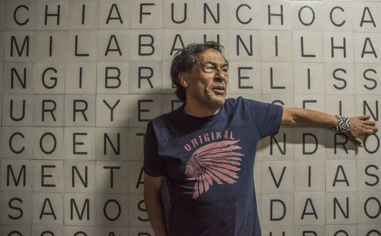 Mężczyzna o ciemnych włosach i karnacji - rdzenny mieszkaniec Brazyli - jedną ręką opera się o ścianę zbudowaną z kostek z literami