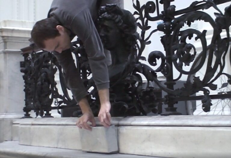 6061b18c49320 Kadr z filmu: młody mężczyzna górną częścią ciała jest przewieszony przez balustradę w holu głównym Zachęty. W rękach trzyma kawałek drewna imitujący marmur