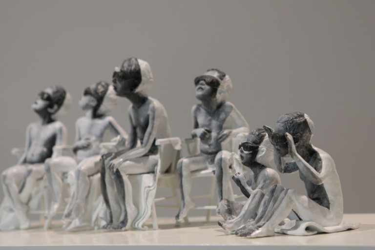 zdjęcie instalacji: szaro-białe figurki przedstawiające nagie dzieci, których oczy przesłonięte są czarnymi goglami. Część dzieci siedzi na krzesłach, część na podłodze