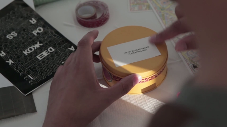 kadr z filmu: widać [dłonie młodej osoby, która nakleja napis na żółte metalowe pudełko