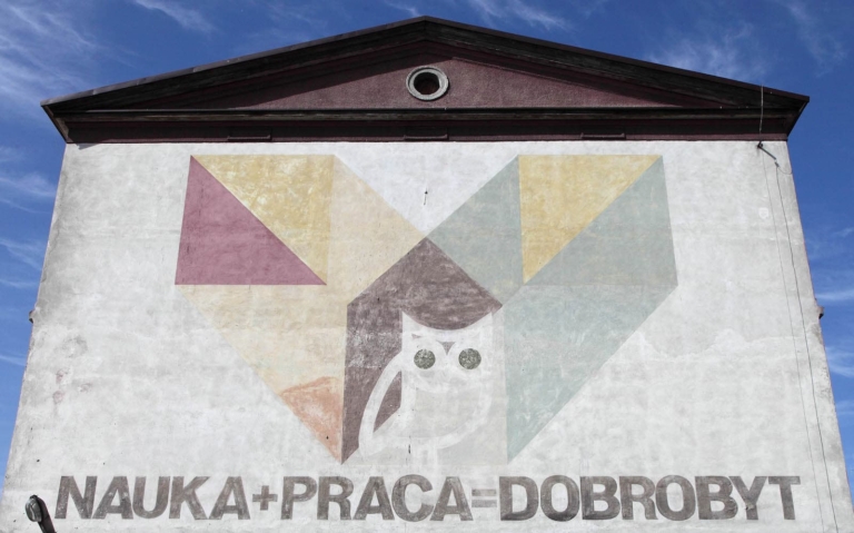 zdjęcie: wypłowiały mural na ścianie budynku. Namalowana biała sowa, oraz kolorowe geometryczne kształty. Poniżej napis: 