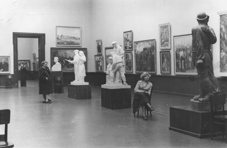 czarno-białe zdjęcie archiwalne: widok sali wystawowej. Na ścianach wiszą obrazy, na środku stoją 3 rzeźby. Obok jednej z rzeźb, na krześle siedzi młoda kobieta