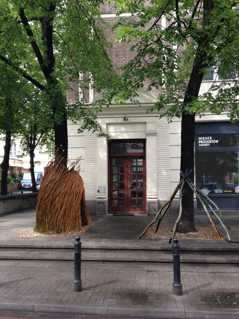 zdjęcie: wejście do budynku mieszkalnego. przed nim, na tarasie, wokół dwóch drzew stoją szałasy z gałęzi i wikliny