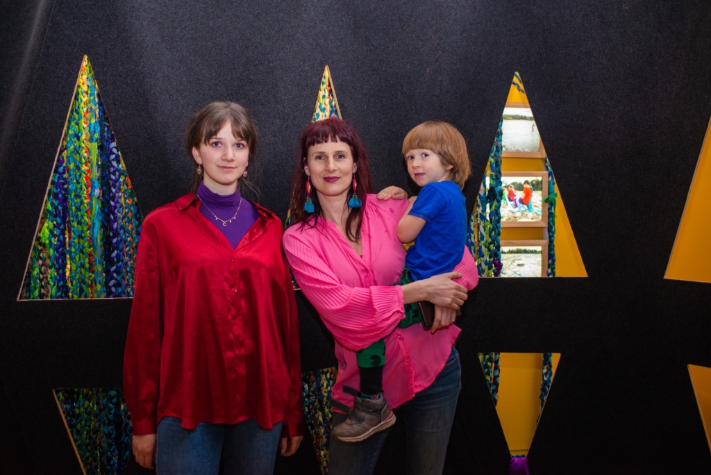 zdjęcie: na tle czarnej ściany z kolorowymi trójkątami stoją dwie kobiety - matka i nastoletnia córka. Matka na rękach trzyma kilkuletniego chłopca