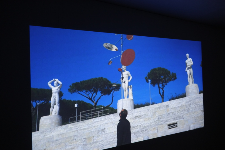 Na pierwszym planie mężczyzna z metalową konstrukcją na głowie. W tle mur, trzy kamienne rzeźby, a w dalszym planie drzewa.