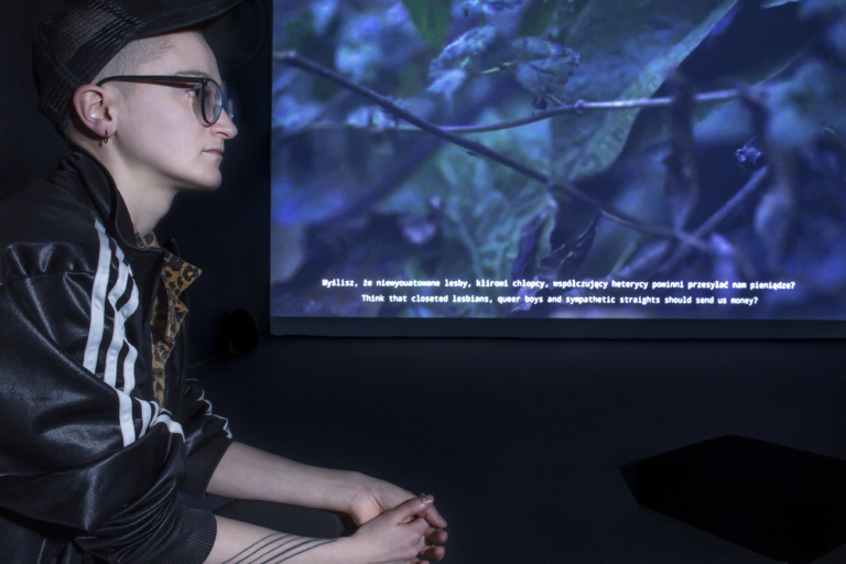 Z lewej strony z profilu artystka w czapce z daszkiem, w okularach i w dresie. Patrzy przed siebie. W tle bardzo duża projekcja jej filmu.