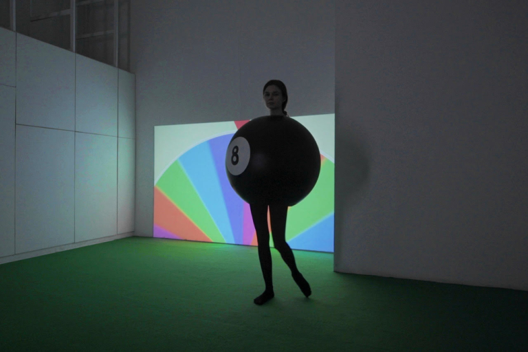 Dziewczyna ubrana w czarną kulę bilardową zakrywającą jej tułów. W tle projekcja wyświetlająca kolorowe koło fortuny.