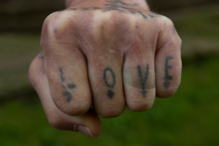 Zdjęcie zaciśniętej pięści. Na każdym palcu wytatuowana rozmazanym tuszem litera. Litery układają się w napis: LOVE.