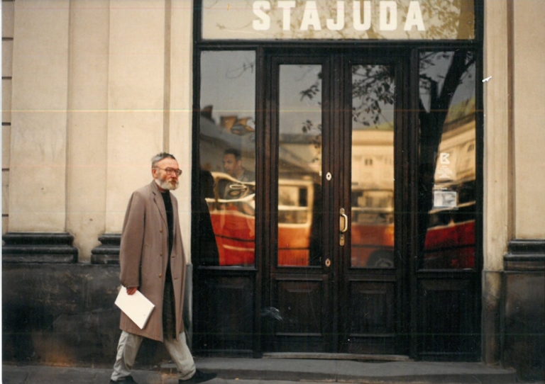 Zdjęcie. Fragment ściany z dużymi przeszklonymi drzwiami, nad nimi napis: Stajuda. Obok drzwi starszy mężczyzna z siwa brodą w płaszczu.