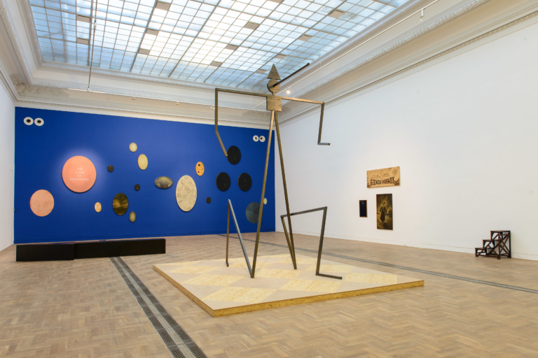 Zdjęcie sali wystawowej. W tle niebieska ściana z okrągłymi i owalnymi obrazami. Na pierwszym planie wysoka patyczakowata postać.