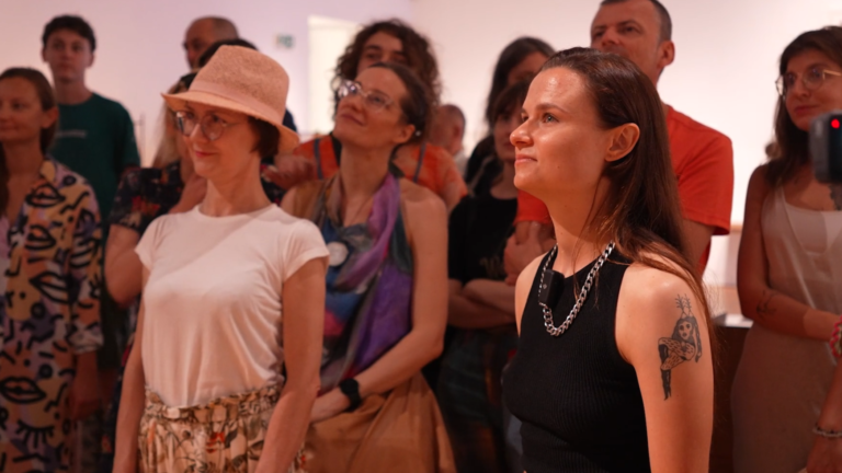 Performatywne oprowadzanie Alki Nauman po wystawie Łzy szczęścia, 30.06.2024, fot. Maria Nitek/archiwum Zachęty Fotografia kolorowa. Grupa osób stojąca blisko i patrząca się przed siebie. Dwie kobiety z przodu uśmiechają się. Kobieta po lewej na głowie ma kapelusz, ta po prawej, na swojej lewej ręce ma tatuaż.