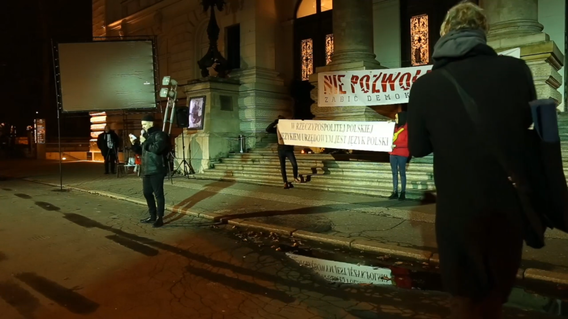 ALT Fotografia kolorowa. Front budynku Zachęty wieczorową porą. Na schodach ludzie z transparentami. Na transparencie napisy „Nie pozwólmy zabić demokracji” na drugim napis „W RZECZPOSPOLITEJ POLSKIEJ JĘZYKIEM URZĘDOWYM JEST JĘZYK POLSKI. Na tle transparentów przemawia mężczyzna, w ręce trzyma mikrofon.