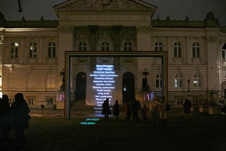 Paweł Żukowski, Napisy końcowe, performans, 30.12.2021, kolekcja Zachęty, fot. Yulia Krivich/archiwum Zachęty Fotografia kolorowa. Front budynku Zachęty wieczorową porą. Rzeźba tęcza, prostokątny kształt w kolorach tęczy. Pod tęczą stoi grupa osób. Na schodach Zachęty stoi duży, biały, kwadratowy ekran, na którym wyświetlane są nazwiska artystów. Imiona i nazwiska wymieniane są pod sobą „Christian Schmidt-Rasmussen, Fin Serck-Hanssen, Ingeborg Strobol, Cuco Suarez, Dermot Seymour, Ernesto Tataflore, Gor Tchahal, Laco Teren, Artur Nacht-Samborski, Andrzej Kurzawski, Włodzimierz Cygan”