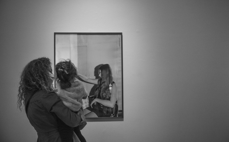 Zdjęcie dokumentujące wystawę w Zachęcie. Kobieta trzyma na rękach małą dziewczynkę. Obie patrzą na fotografię innej kobiety, która wyciąga rękę. Zdjęcie jest tak zrobione, że mamy wrażenie, że osoba z fotografii dotyka kobiety oglądające prace na wystawie