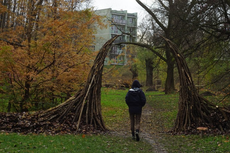 opis alt: Zdjęcie parku jesienią. Przez bramę z gałęzi i patyków przechodzi w głąb parku postać ludzka widziana od tyłu.