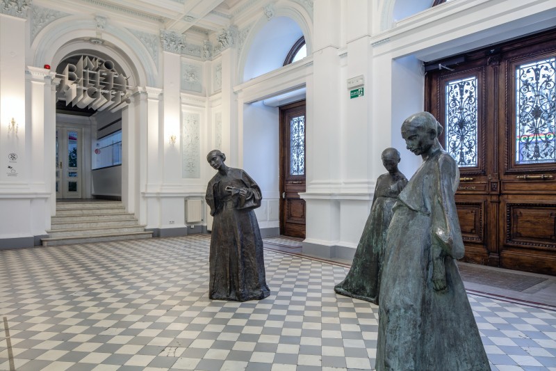 Zdjęcie: w holu głównym Zachęty, przed historycznymi, drewnianymi drzwiami galerii stoją trzy rzeźby z brązu Xawerego Dunikowskiego. Przedstawiają trzy brzemienne kobiety