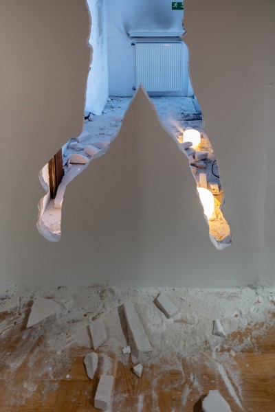 Zdjęcie fragmentu ekspozycji "Rzeźba w poszukiwaniu miejsca". U góry dziura w ścianie w kształcie tułowia z nogami. Na podłodze fragmenty tynku i gipsu. Po drugiej stronie dziury fragment klatki schodowej. 
