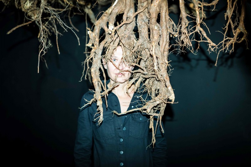 zdjęcie: kobieta w krótkich włosach, w czarnej koszuli stoi w ciemnym pomieszczeniu. Z sufitu zwisają karpy drzew z korzeniami. Twarz kobiety jest ledwo widoczna zza korzeni