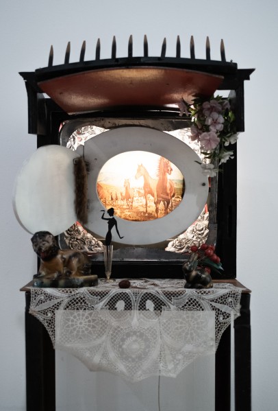 Na stole przykrytym serwetką stoi "telewizor", którego ekran wpisany jest w deskę sedesową. Zwieńczenie w formie grabi, wokół kwiatki i figurki.