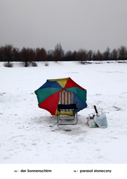 na zdjęciu, teren zasypany śniegiem, w górnej części szare niebo i drzewa, na środku turystyczne krzesełko i kolorowy plażowy parasol. Obok krzesła wiaderko i biała plastikowa torba.