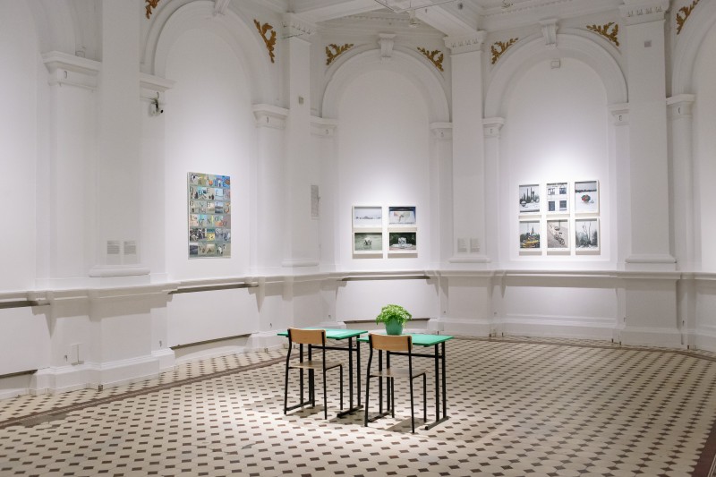 Zdjęcie sali ekspozycyjnej. Na ścianach wiszą dzieła sztuki - oprawione fotografie i grafika. Na środku sali stoją dwie zielone ławki szkolne z krzesłami. Na jednej z ławek stoi roślina. 