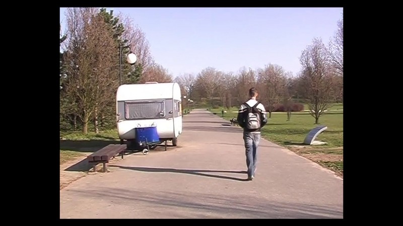 Stopklatka z wideo. Widok parku wczesną wiosną. Asfaltowa alejka, po lewej stronie przyczepa kempingowa, po prawej, w głąb kadru idzie chłopak z plecakiem na plecach.