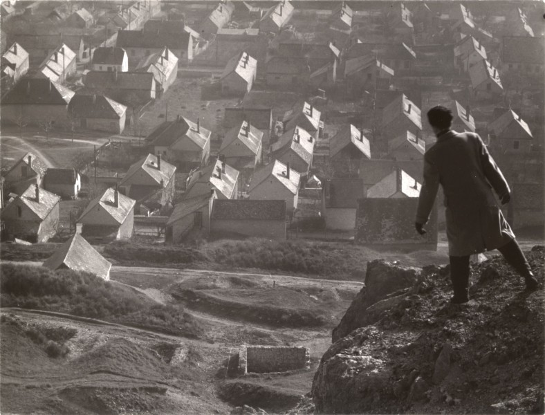 czarno-białe zdjęcie: na pierwszym planie mężczyzna stojący na skale, patrzący w dół. W oddali, w dolinie, miasteczko pełne drewnianych domów